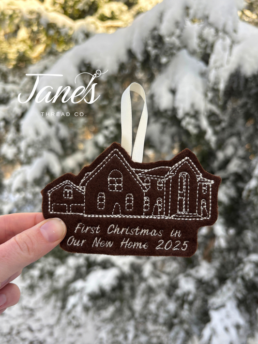 Decorative house ornament with 'First Christmas in Our New Home 2025' text, held against a snowy tree background.