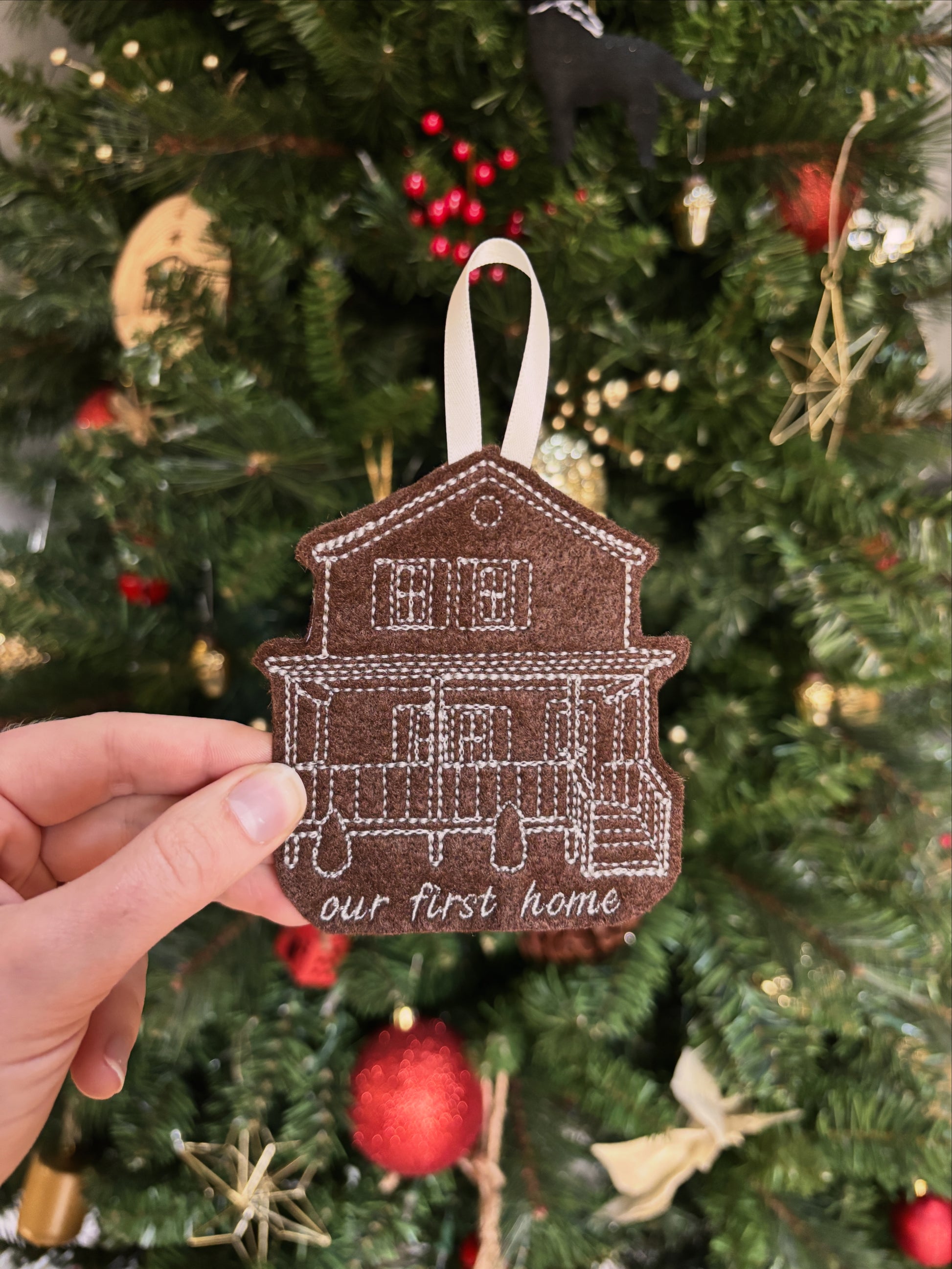 Decorative house ornament with 'our first home' text held in front of a Christmas tree.