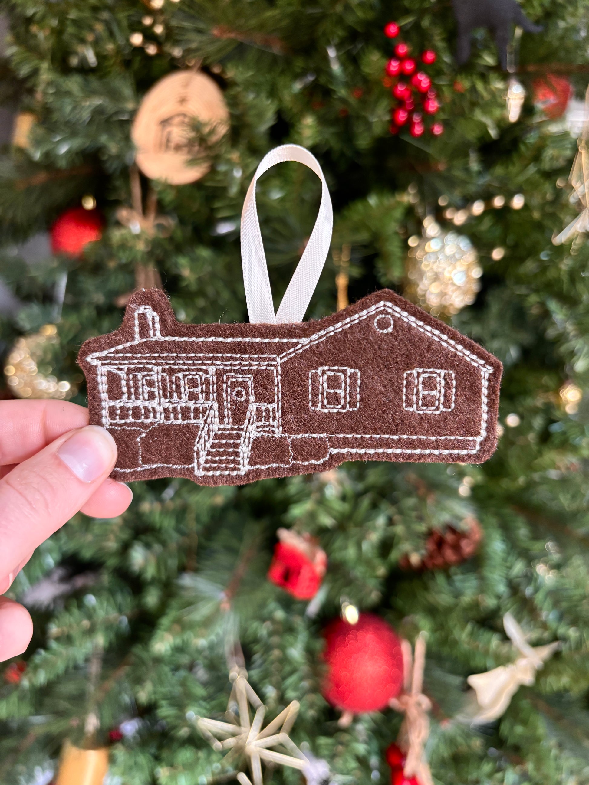 Hand holding a house-shaped ornament in front of a decorated Christmas tree.