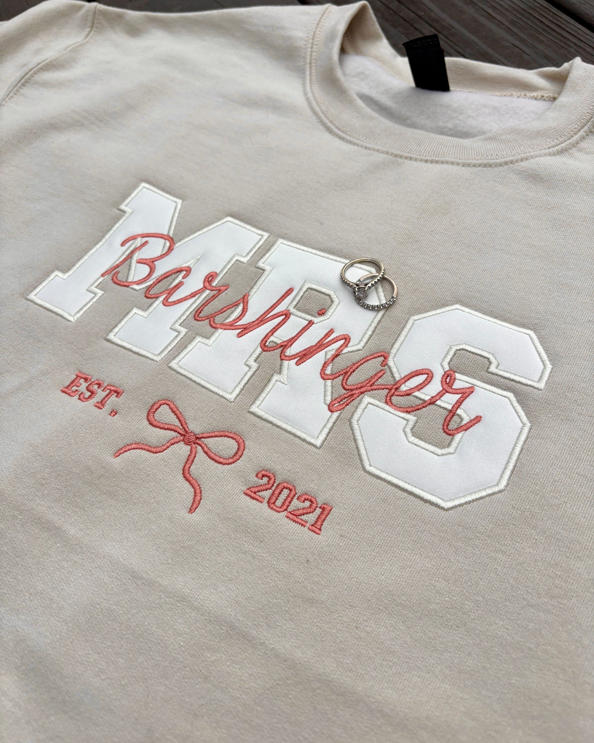 Beige sweatshirt with 'MRS. Barshinger' and 'EST. 2021' text on a wooden surface