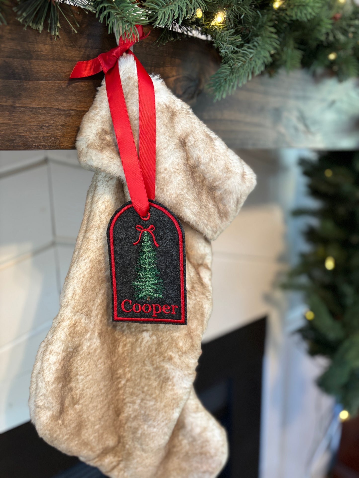 Beige Christmas stocking with a red ribbon and 'Cooper' tag hanging on a fireplace mantle.