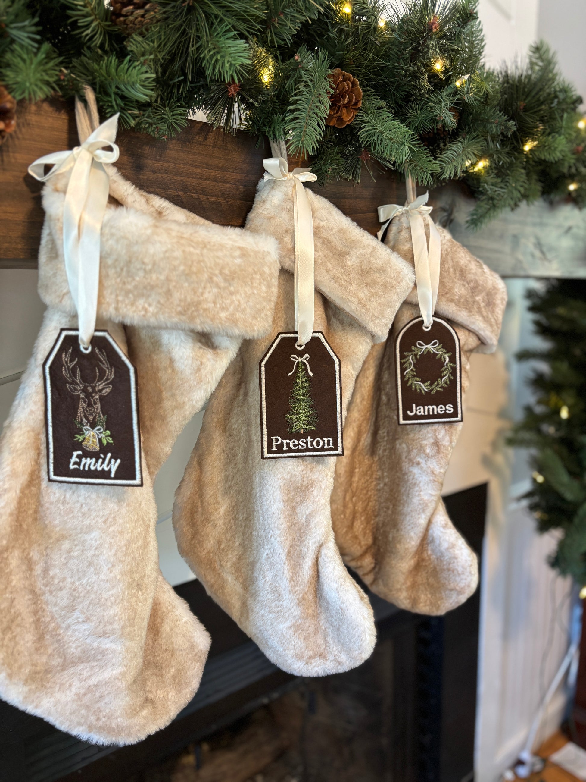 Three beige Christmas stockings hanging on a fireplace mantle with decorative tags.