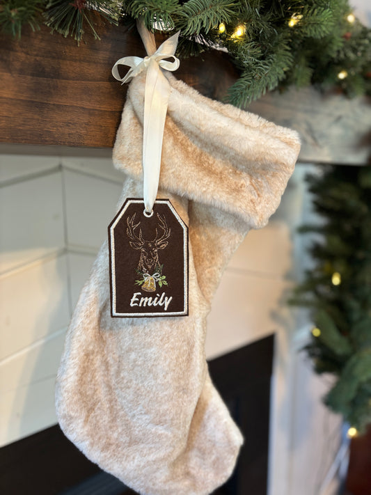Beige Christmas stocking with a name tag hanging under a decorated tree.
